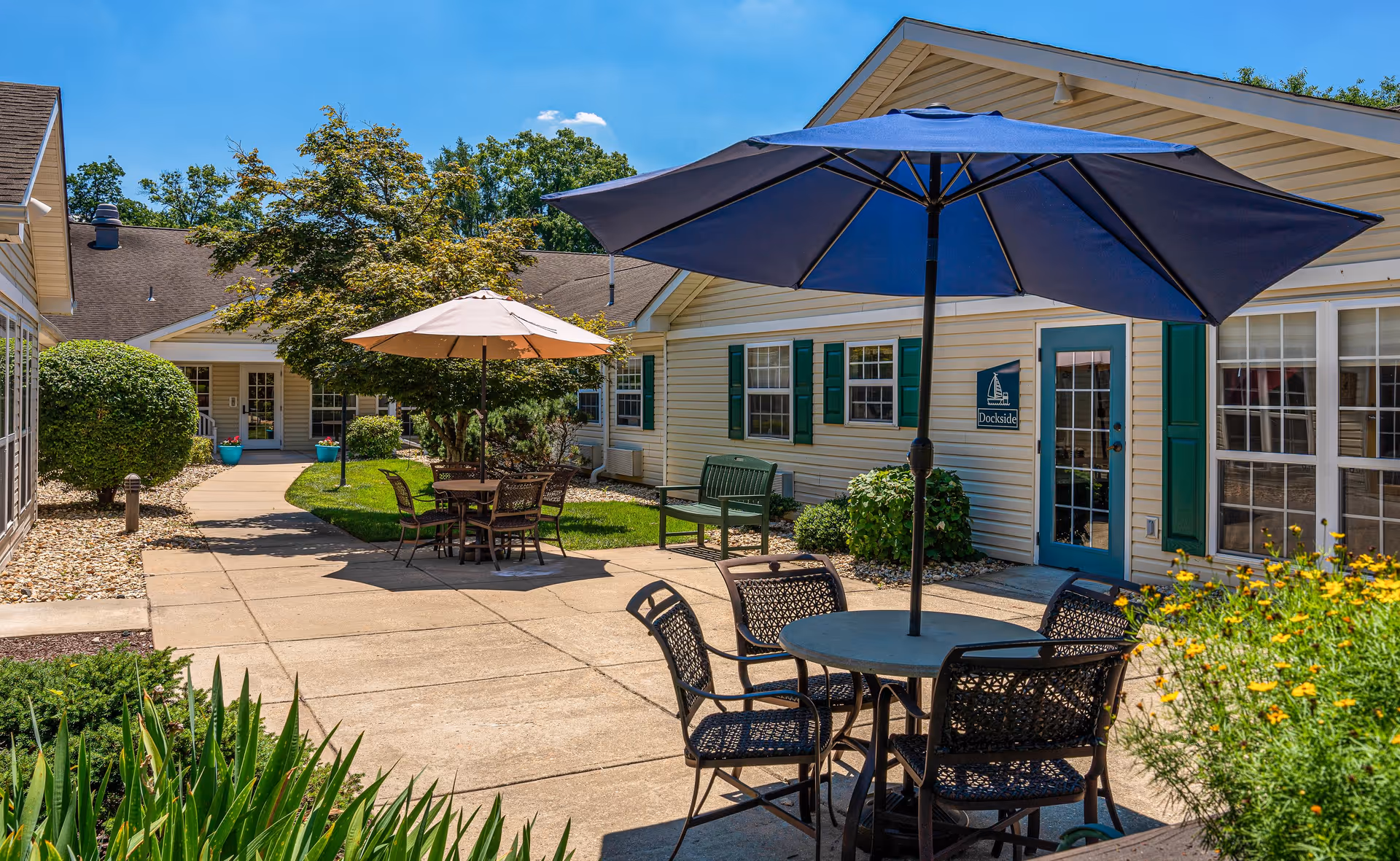Outdoor courtyard area at Arden Courts - ProMedica Memory Care Community (Kensington) featuring patio tables with umbrellas, chairs, green bushes, trees, and beige buildings with green shutters under a clear blue sky.