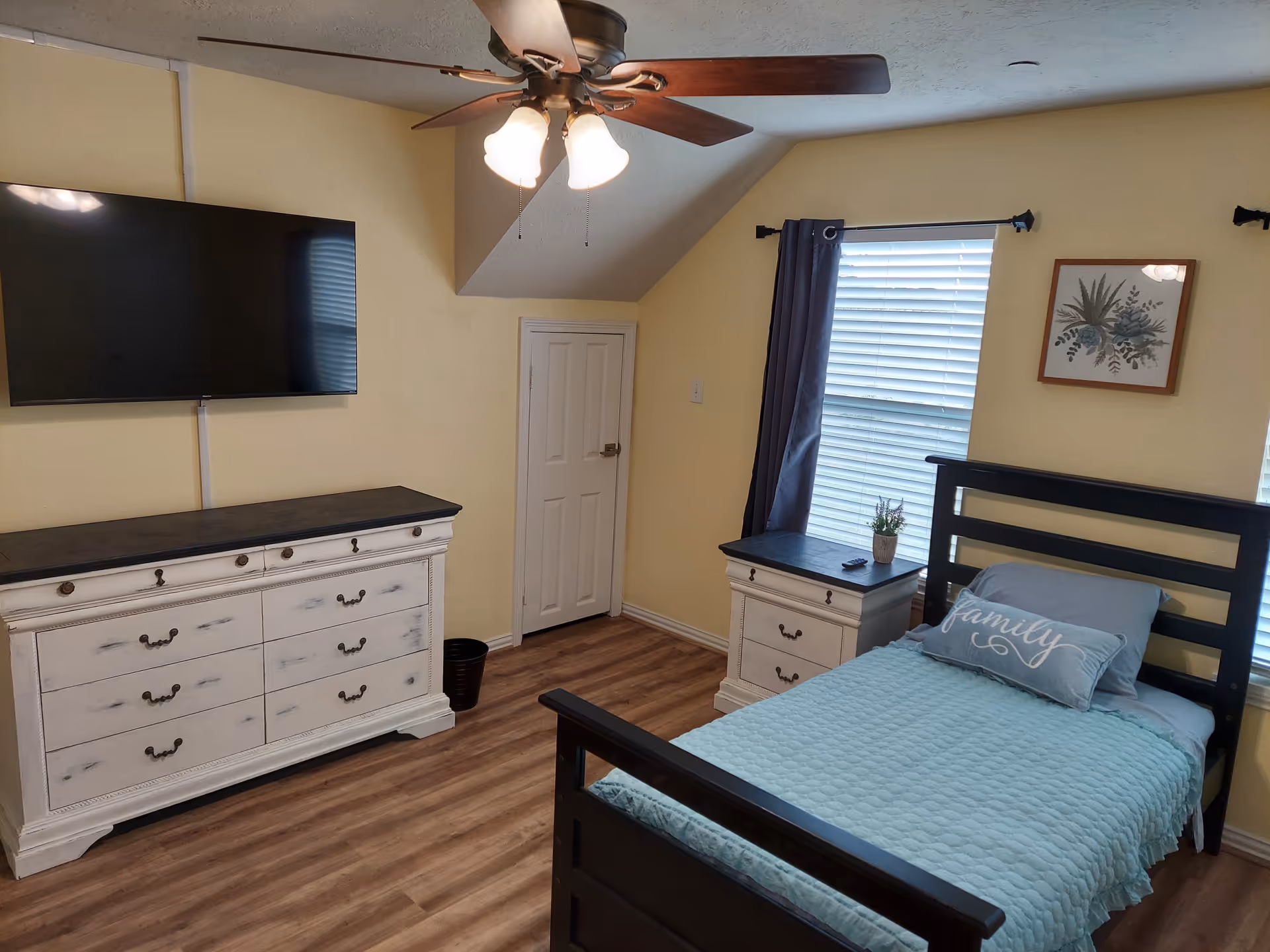 A cozy bedroom with a black wooden bed frame, a light blue quilt and pillows including one with the word 'family'. There is a white dresser with a dark top beneath a mounted flat-screen TV, a matching nightstand with a small plant, a window with blinds and dark curtains, a ceiling fan with lights, and a small white door in the corner.