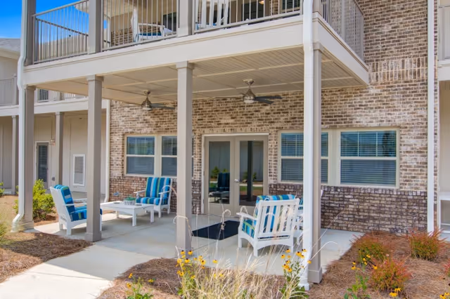 Outdoor patio area at a senior living facility with cushioned chairs and a coffee table under a covered porch. The building has brick walls, two ceiling fans, and windows with blinds. There are plants and flowers in the landscaped area around the patio.