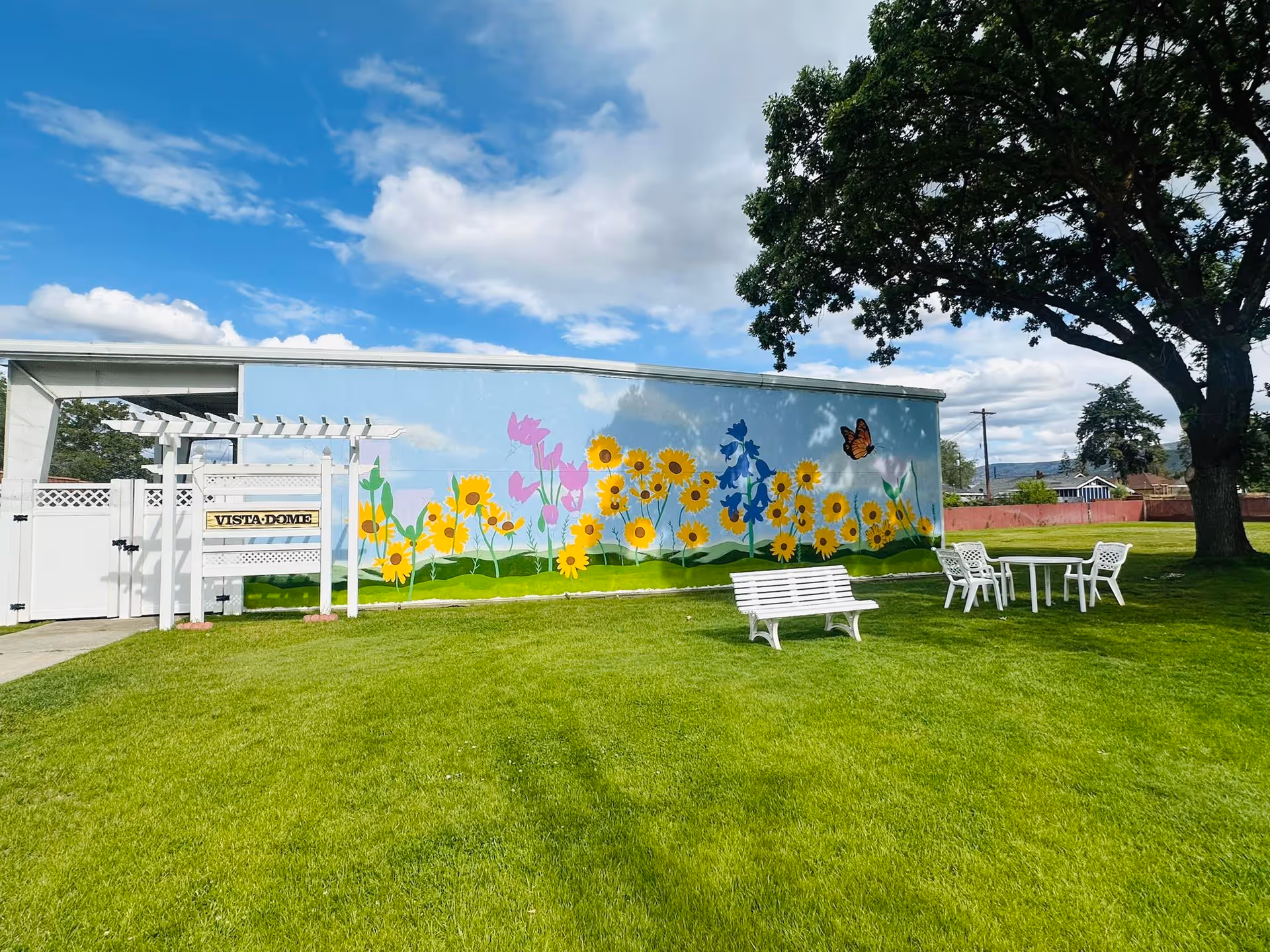 Outdoor grassy area with white benches and chairs around a table, a large tree on the right, and a building wall painted with a colorful mural of sunflowers, other flowers, and a butterfly under a partly cloudy blue sky.