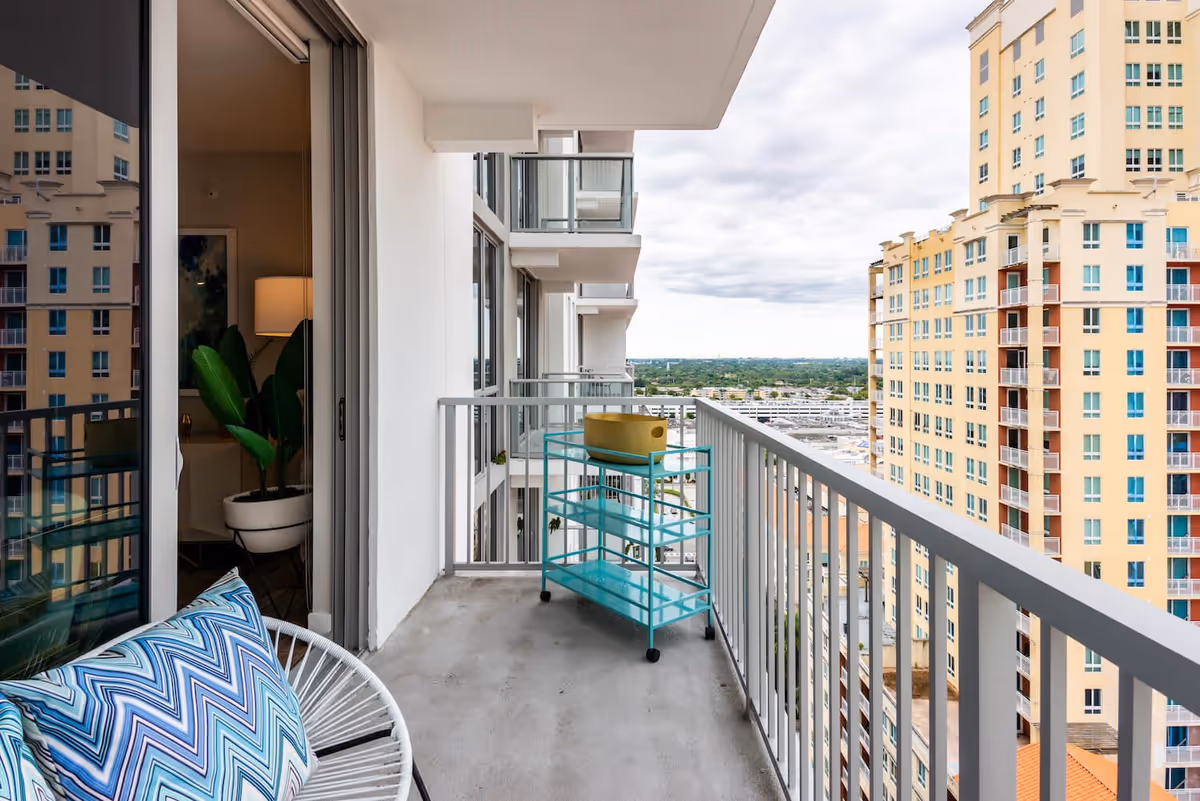 Balcony of a high-rise apartment with a white metal chair featuring a blue and white chevron-patterned cushion, a turquoise rolling cart with a yellow bowl on top, and a view of nearby tall buildings and a distant landscape under a cloudy sky.