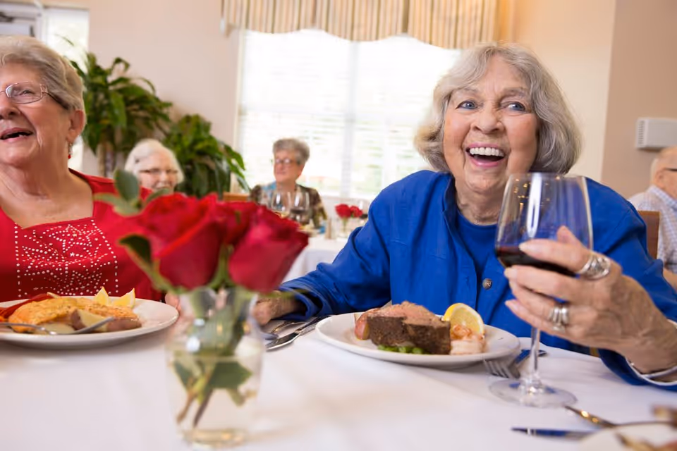 Elderly women smiling and enjoying plated meals and wine at a communal dining table with a vase of red roses.