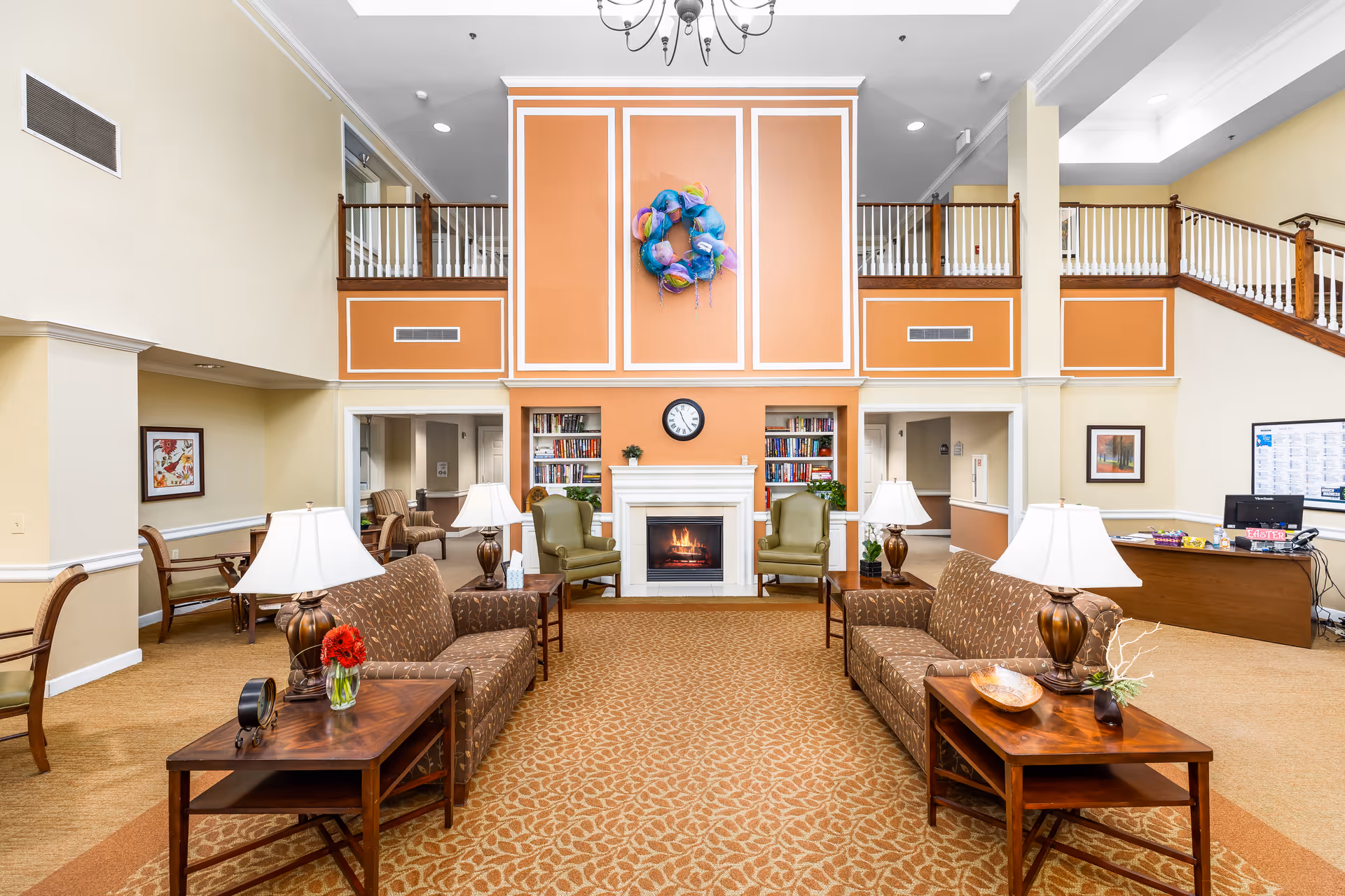 A spacious senior living facility common area with two brown patterned sofas facing each other, each with a wooden side table holding a lamp and decorative items. In the center background is a white fireplace with a clock above it, flanked by bookshelves and two green armchairs. The walls are painted in warm beige and orange tones, with a colorful wreath hanging above the fireplace. A staircase with wooden railings leads to an upper level balcony overlooking the room.