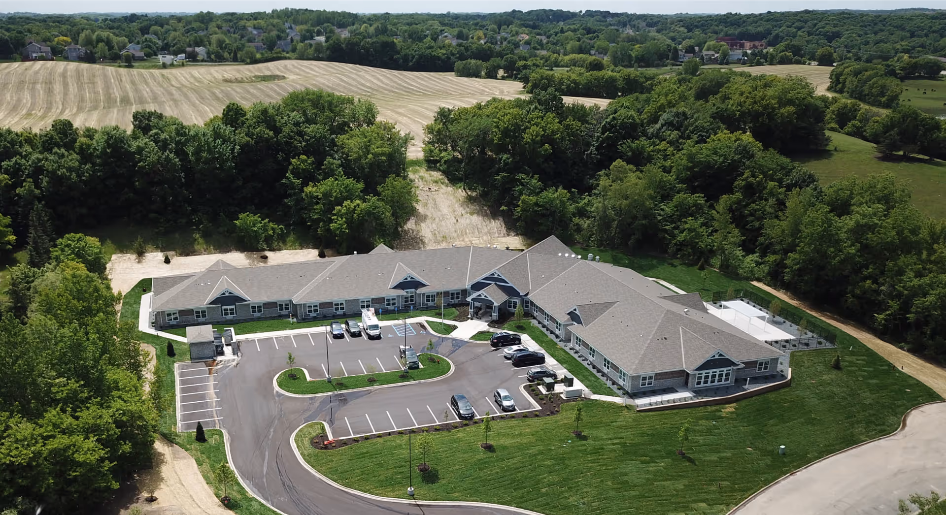 Aerial view of a single-story senior living building with a circular driveway, parking lot, lawns, and surrounding trees and fields.