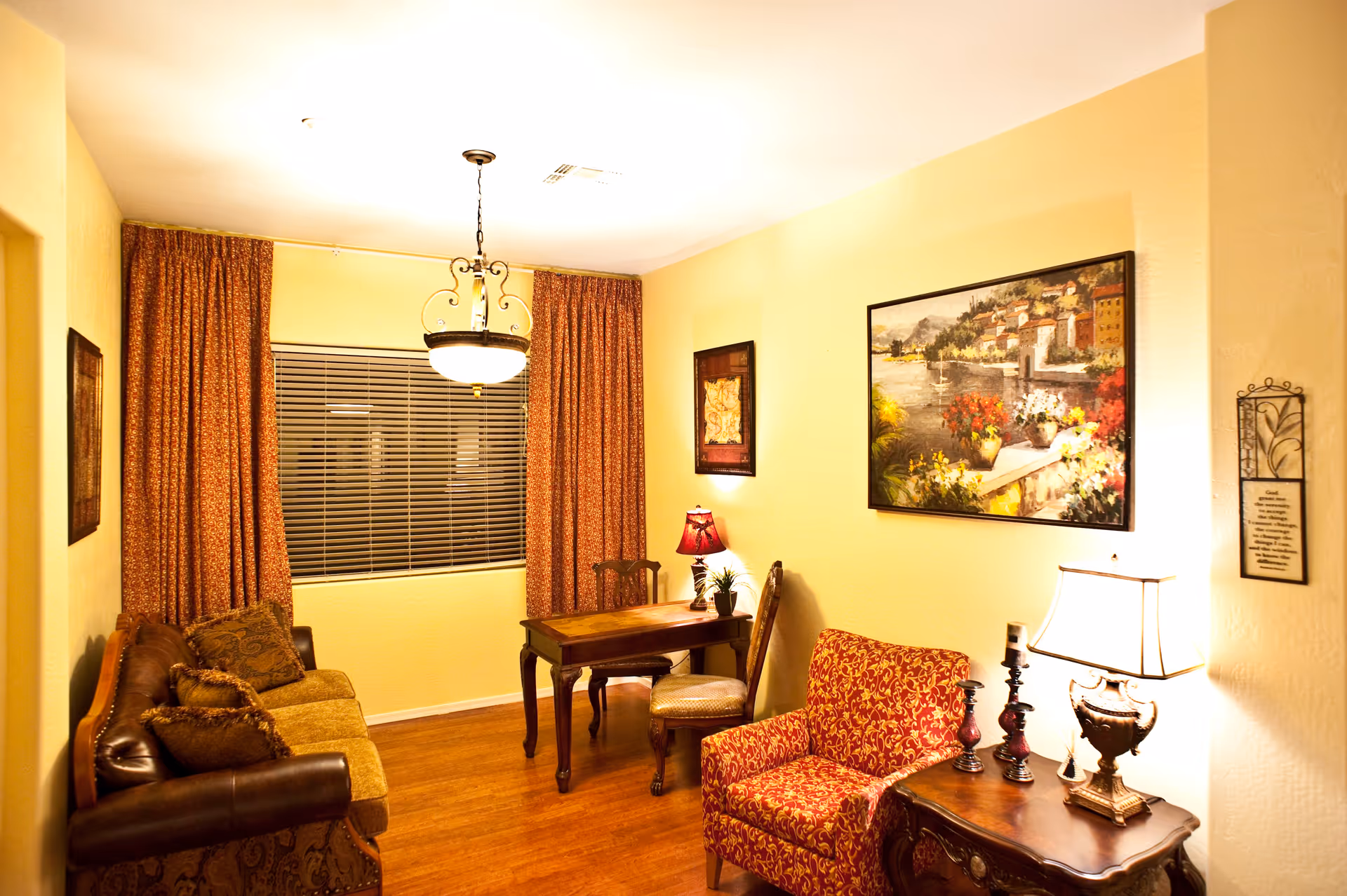 A cozy living room with a brown leather and fabric sofa on the left, a wooden table with two chairs in the center, and a red patterned armchair next to a wooden side table with two lamps on the right. The walls are painted light yellow and decorated with framed paintings and a wall hanging. The window has closed blinds and orange patterned curtains.