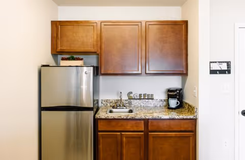 Small kitchenette with a stainless steel refrigerator on the left, wooden cabinets above and below a granite countertop. The countertop has a small sink, a coffee maker with a white mug, and decorative letters spelling 'COFFEE'.