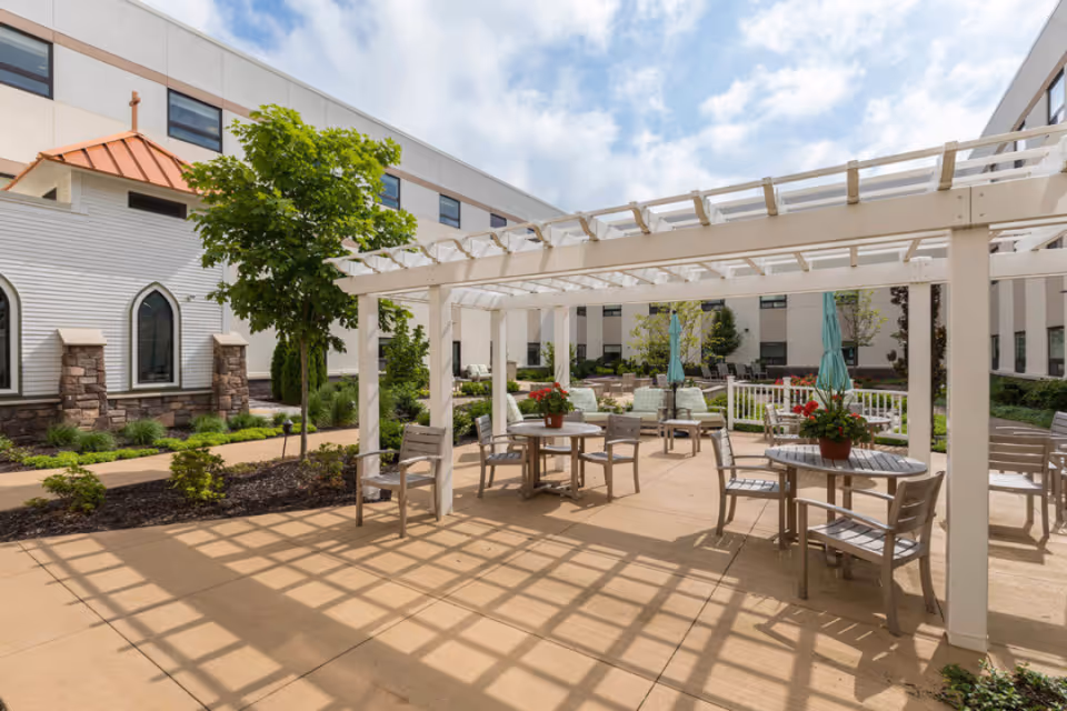 Sunlit courtyard with a white pergola, round tables and chairs, potted flowers, and the surrounding building.
