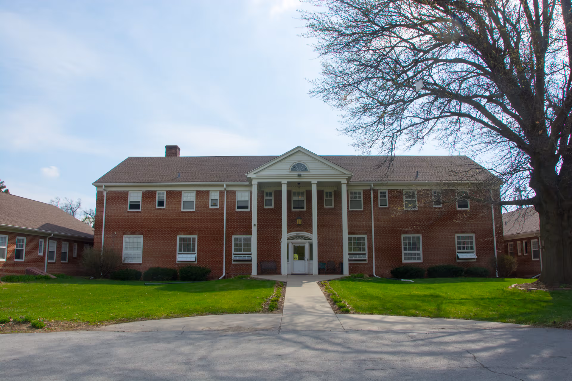 Front exterior view of a two-story brick building with white trim and columns at the entrance, surrounded by green grass and a large tree on the right side under a partly cloudy sky.
