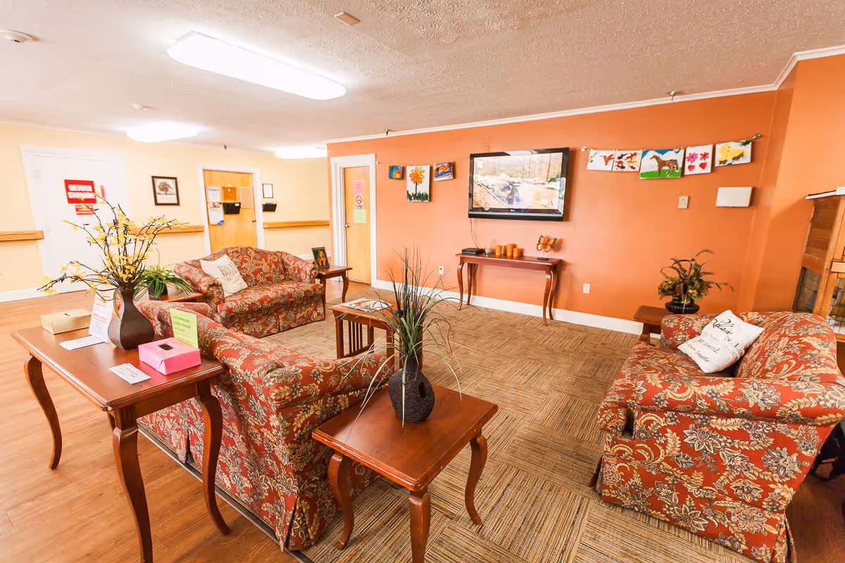 A cozy living room area in Oakview Nursing and Rehabilitation Center featuring three patterned red sofas arranged around a central wooden coffee table with a plant. There are additional wooden side tables with decorative plants and items. A flat-screen TV is mounted on an orange accent wall, with colorful artwork hanging on a string above it. The room has a carpeted area and wood flooring, with bright overhead lighting and doors leading to other parts of the facility.