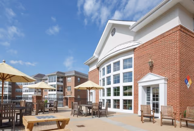 Outdoor patio area at Matthews Glen with several wooden tables and chairs under large beige umbrellas. The patio is adjacent to a large brick building with tall windows and a white door. Another multi-story brick building is visible in the background under a partly cloudy blue sky.