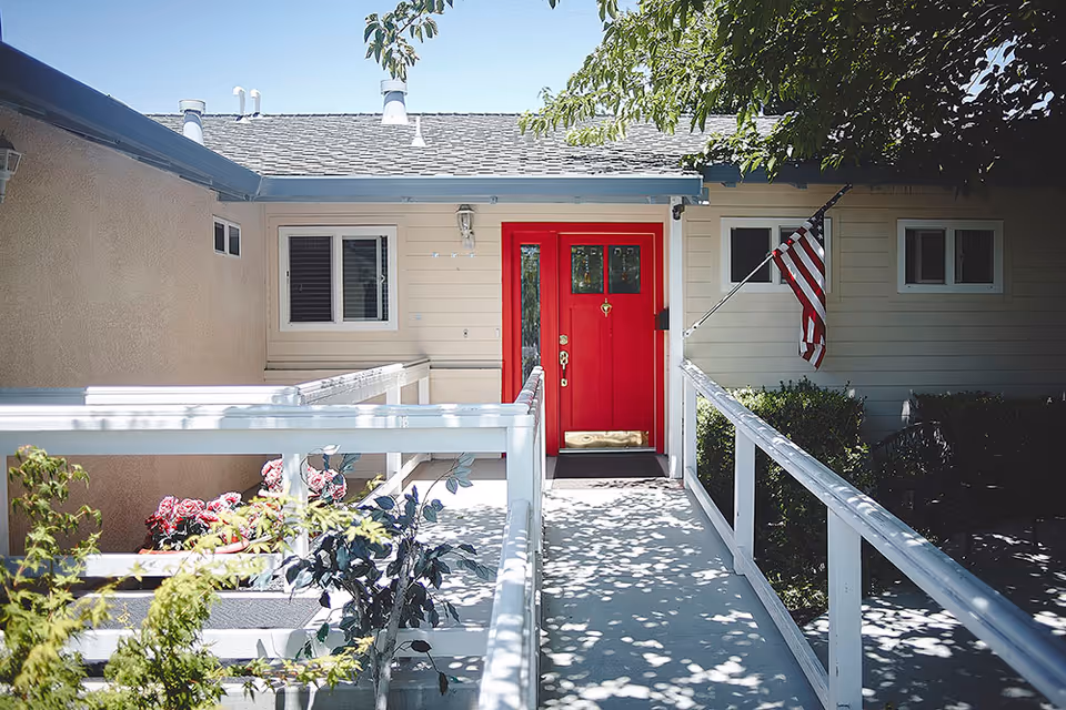 Entrance to a care home with a bright red door, white railings along a ramp, beige exterior walls, an American flag mounted on the right side, and some greenery and flowers around the walkway.