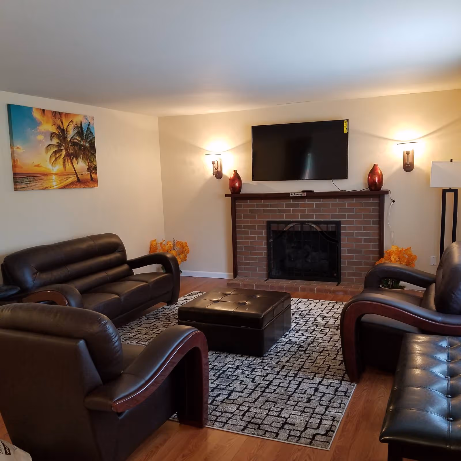 A cozy living room with brown leather sofas and armchairs arranged around a black and white patterned rug. A brick fireplace with a black screen is centered on the far wall, above which a flat-screen TV is mounted. Two red decorative vases sit on the fireplace mantel, flanked by wall sconces emitting warm light. A floor lamp stands to the right, and a colorful painting of a beach sunset with palm trees hangs on the left wall.