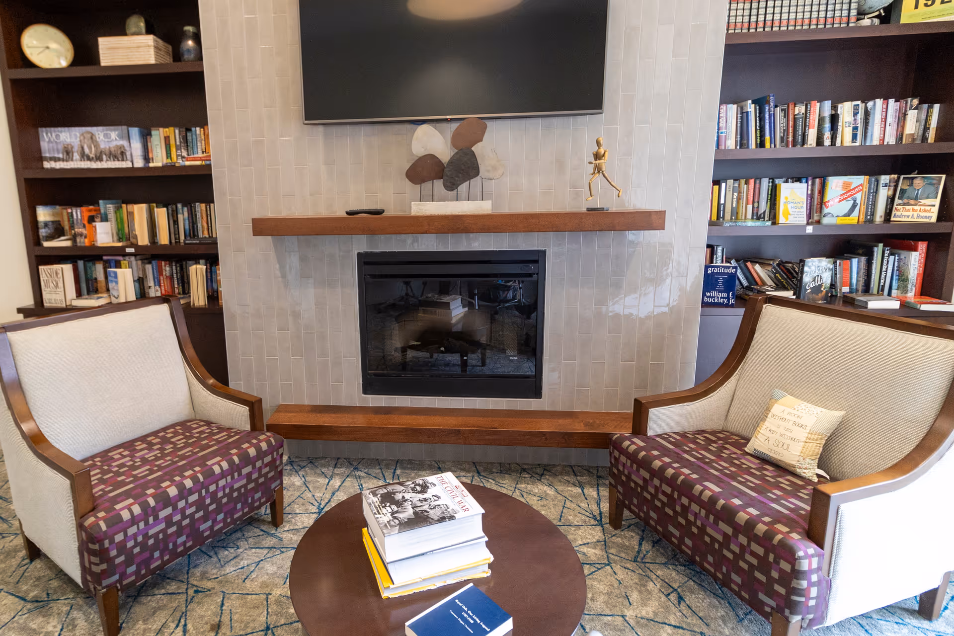 A cozy sitting area in a library or lounge with two upholstered armchairs facing a round wooden table stacked with books. Behind the chairs is a modern fireplace with a wooden mantel, above which is a flat-screen TV. On either side of the fireplace are built-in bookshelves filled with books and decorative items.