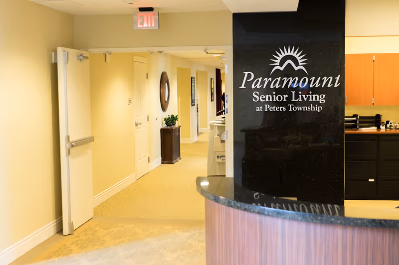 Reception area with a curved desk and a black pillar sign reading "Paramount Senior Living at Peters Township" beside a hallway.