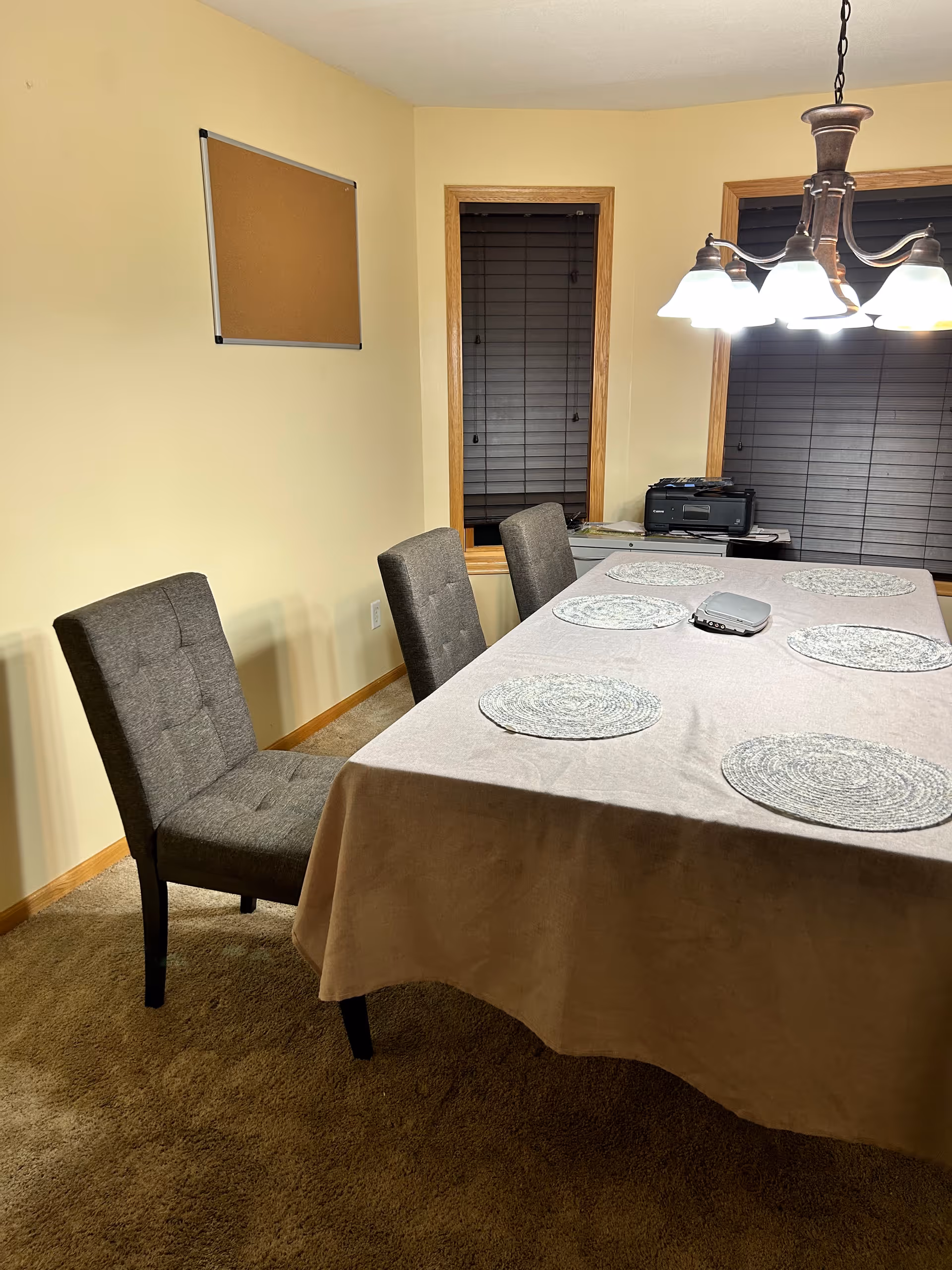 A dining room with a rectangular table covered with a beige tablecloth and six round woven placemats. There are three gray upholstered chairs around the table. Two windows with closed dark blinds are visible in the background, along with a printer on a small table or desk. A hanging light fixture with multiple bulbs illuminates the room. A cork bulletin board is mounted on the wall.