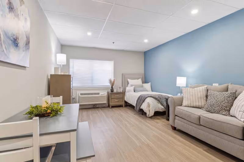 A bright and clean senior living room featuring a single bed with white bedding and gray throw, a beige sofa with multiple cushions, a wooden nightstand with a lamp, a window with blinds, a small dining table with chairs, and light wood flooring. The walls are painted in light gray and blue tones, and there is a decorative painting on the wall.