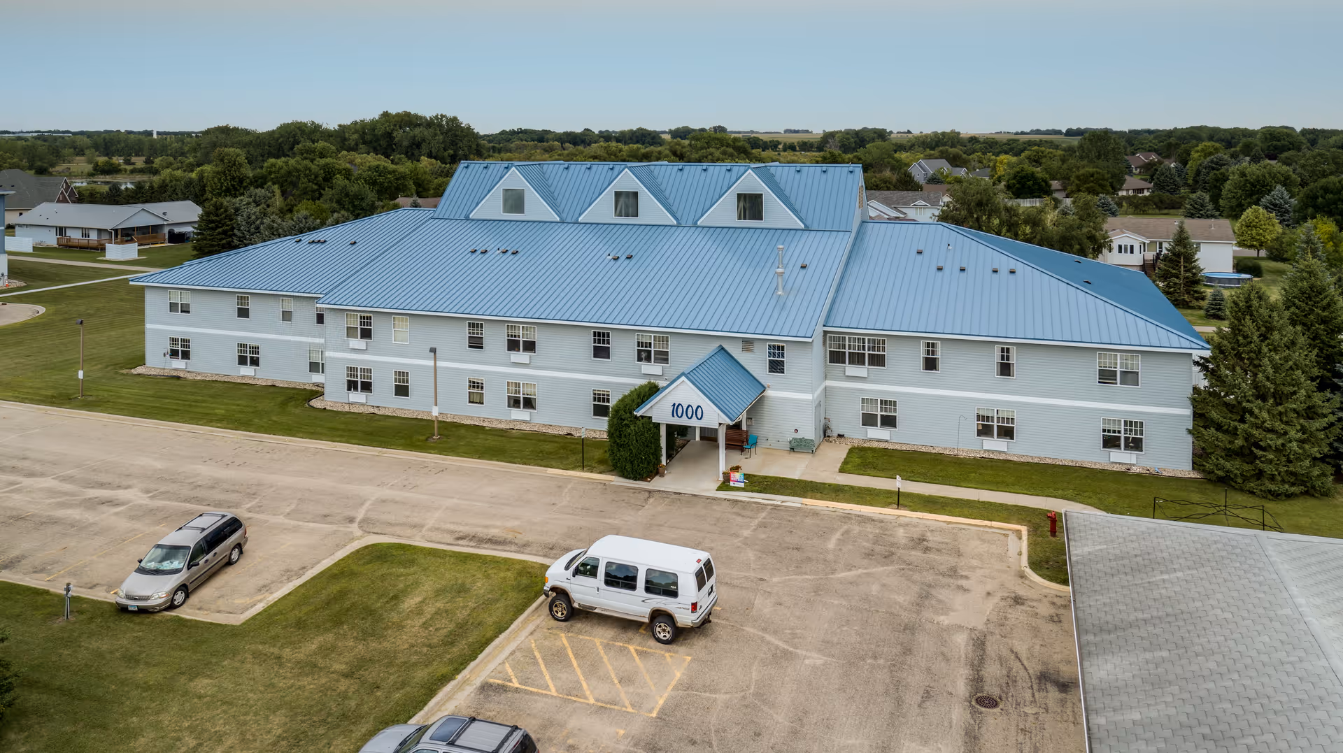 Aerial view of a two-story light-blue senior living building with a blue metal roof, entrance marked '1000', and a parking lot with a van.