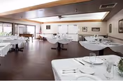 Spacious dining room with multiple round and rectangular tables draped in white tablecloths and set with plates and glasses, a piano and windows along the back wall.