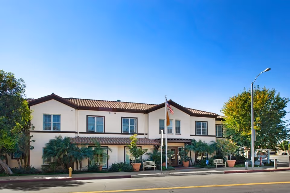Exterior view of a two-story senior living facility building with a tiled roof, multiple windows, and an entrance with benches and potted plants. There are two flagpoles with flags in front of the building, and a street with a sidewalk and a streetlamp is visible in the foreground under a clear blue sky.