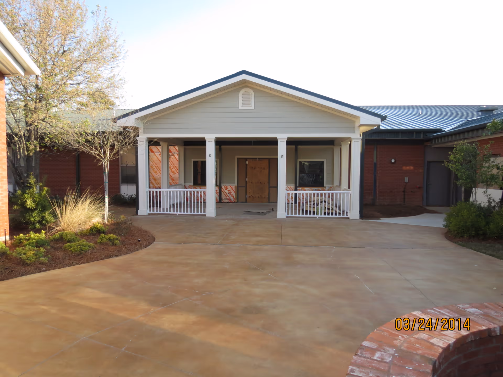 Exterior view of a building with a covered porch supported by white columns and a concrete driveway in front. There are small landscaped areas with plants and trees on either side of the driveway. The building has a light gray facade with a dark roof and red brick walls on the sides.