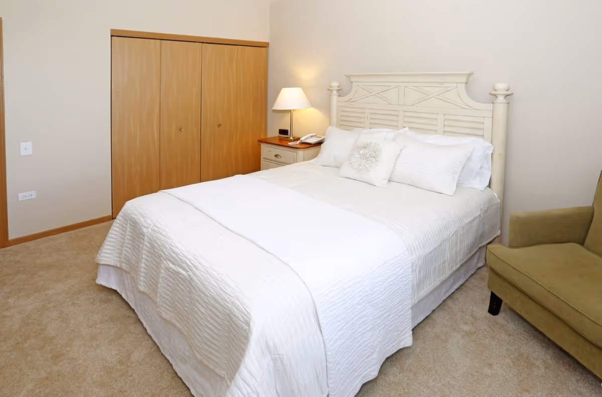 A tidy bedroom with a white-made bed, decorative headboard, nightstand and lamp, wooden closet doors, and an olive armchair.