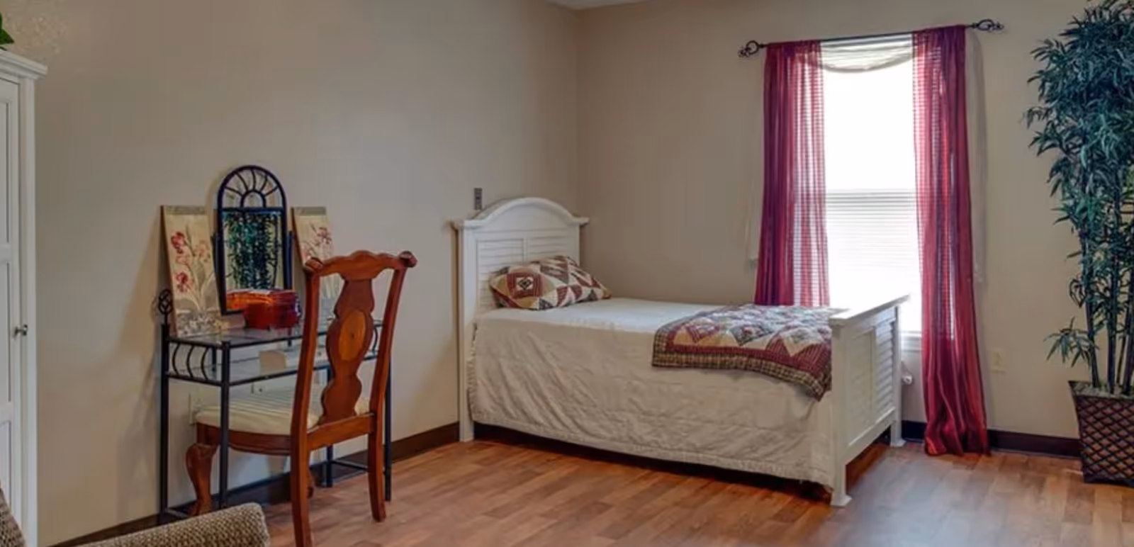 A bedroom with a single white bed featuring a quilt and pillow, a wooden chair, a glass-top vanity table with decorative panels and a mirror, a window with red curtains, and a tall potted plant in the corner.