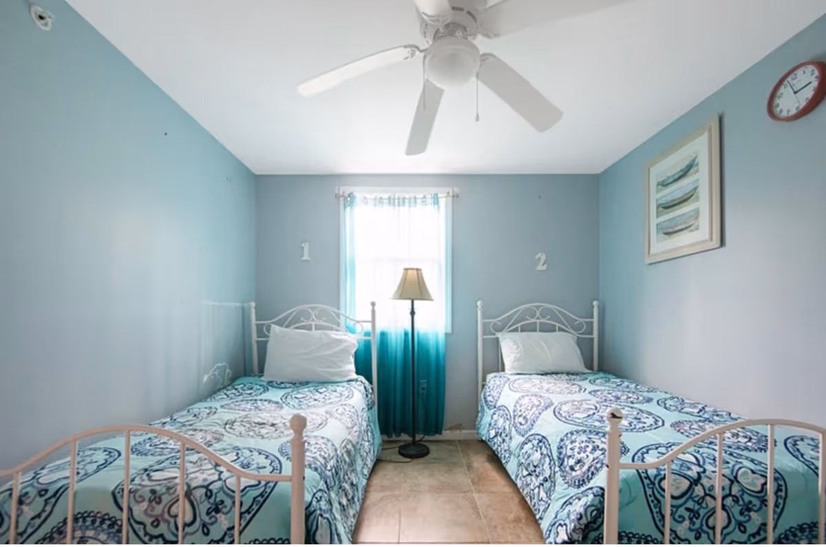 A small bedroom with two single beds placed parallel to each other against opposite walls. Each bed has a white metal frame and is covered with a blue patterned bedspread. A window with blue curtains is centered on the back wall between the beds, and a floor lamp stands in front of the window. The walls are painted light blue, and there is a ceiling fan above. A framed picture of boats and a round wall clock are visible on the right wall.
