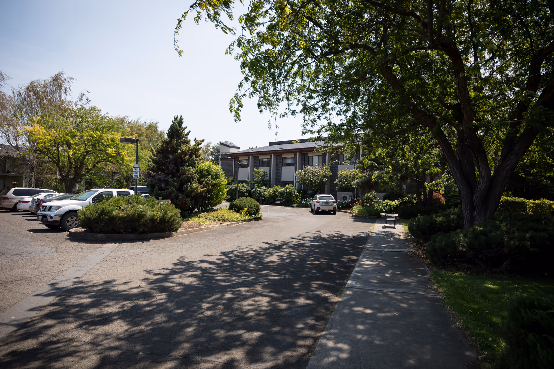 View of the exterior of a senior living facility named Cherry Heights Living, showing a parking area with several cars, surrounded by trees and greenery under a clear sky.