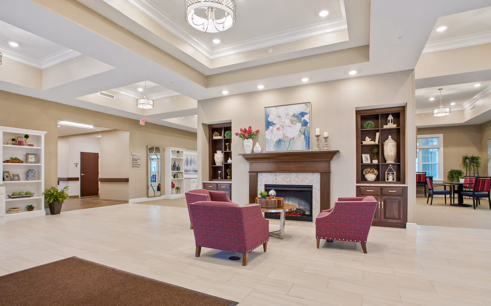 A spacious and well-lit senior living facility common area with a fireplace centered between two built-in wooden shelves decorated with vases, plants, and candles. In front of the fireplace are three red patterned armchairs arranged around a small round wooden table. The room has light-colored tiled flooring, beige walls, and a tray ceiling with recessed lighting and chandeliers. To the right, there is a dining area with tables and chairs, and to the left, a hallway leading to other parts of the facility.