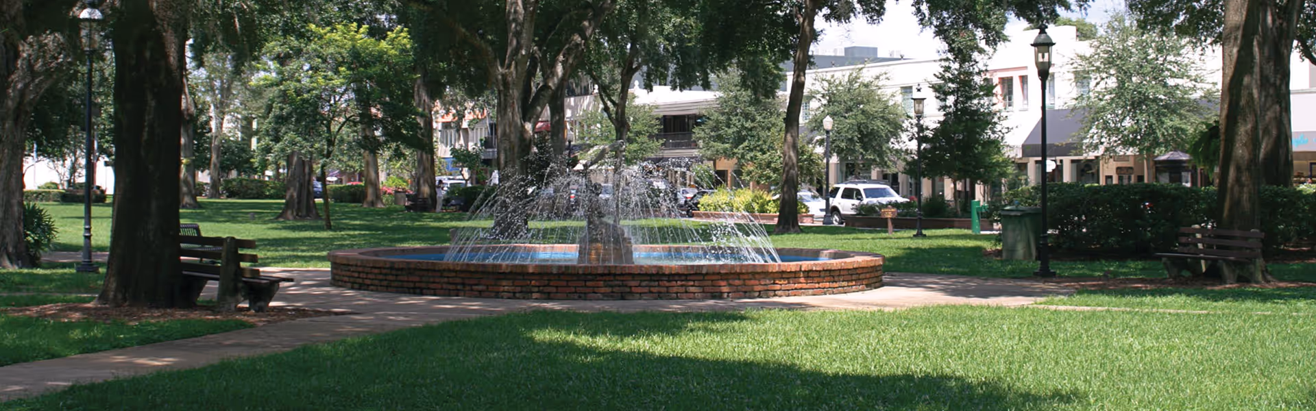A brick circular fountain in a shaded park surrounded by trees, benches, grassy lawn, and buildings in the background.