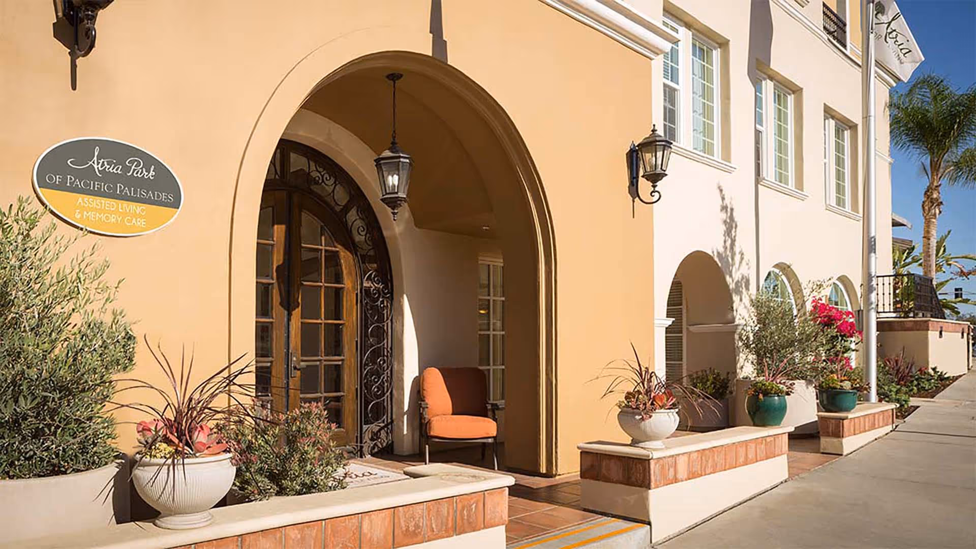 Exterior view of Atria Park of Pacific Palisades building entrance with arched doorway, potted plants, outdoor seating, and a flagpole with the facility's flag under a clear blue sky.