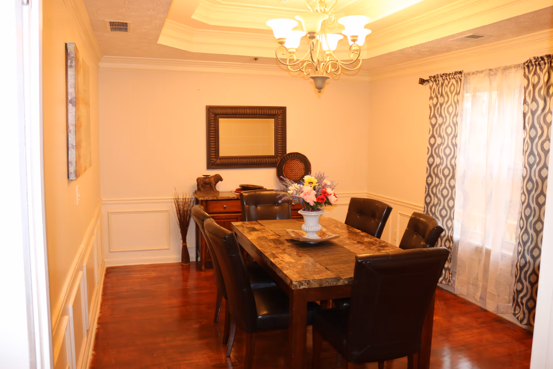 Dining room with a wooden table surrounded by six leather chairs, a chandelier overhead, and a vase of flowers centerpiece.