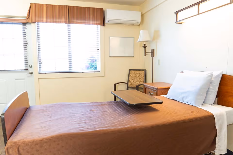 A simple senior facility bedroom with a single bed, bedside table, chair, and a window with blinds.