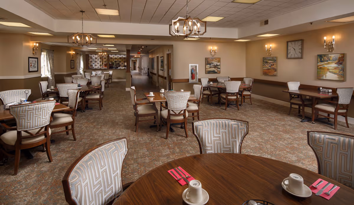 A spacious dining room with multiple round and square wooden tables surrounded by cushioned chairs with patterned upholstery. The room has beige walls adorned with framed paintings and wall sconces. There are chandeliers hanging from the ceiling providing warm lighting. The carpet has a subtle floral pattern, and the tables are set with cups, saucers, and pink napkins with silverware.