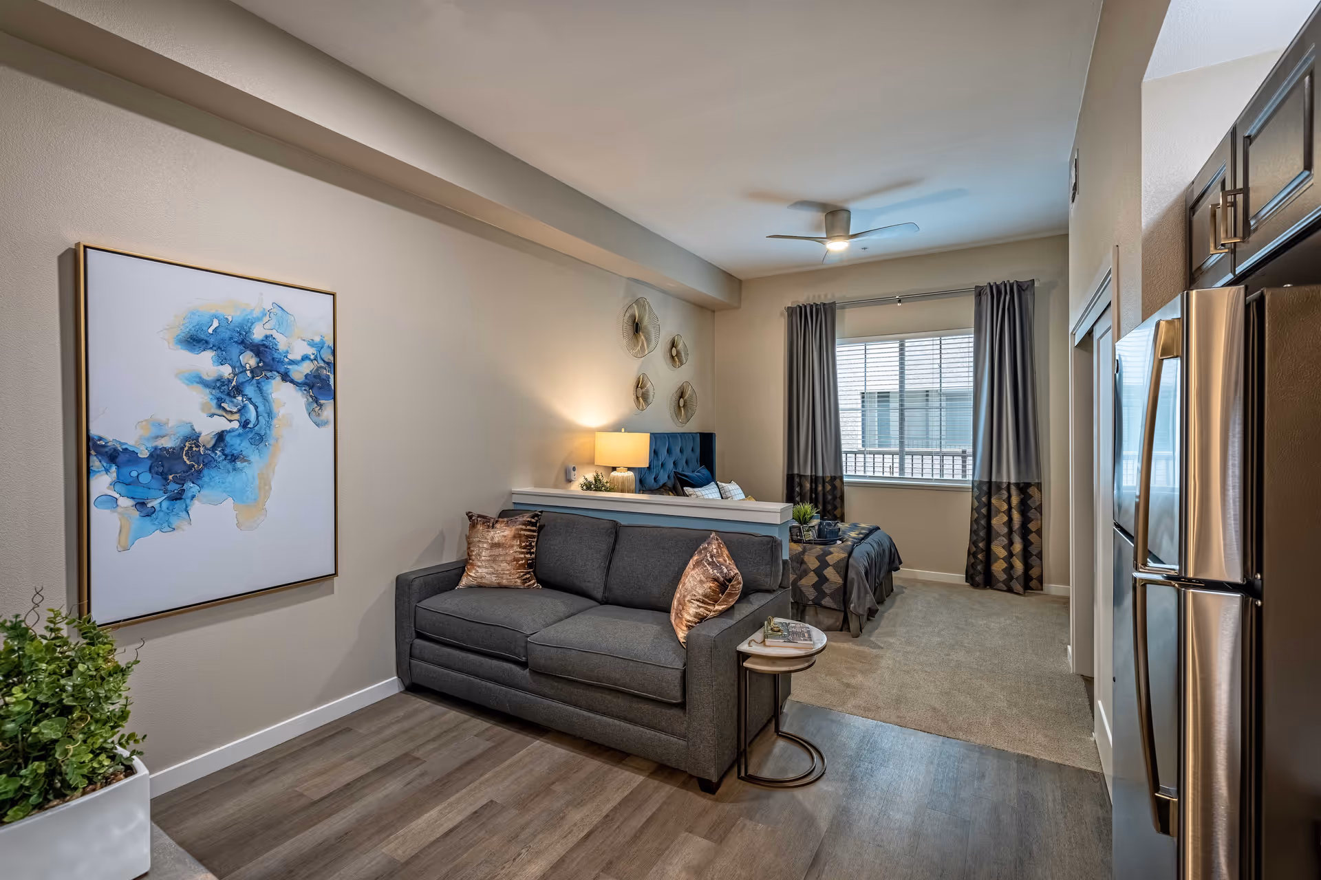 Furnished studio interior with a gray sofa and side table in front of a bed area, window, artwork, and a stainless steel refrigerator.