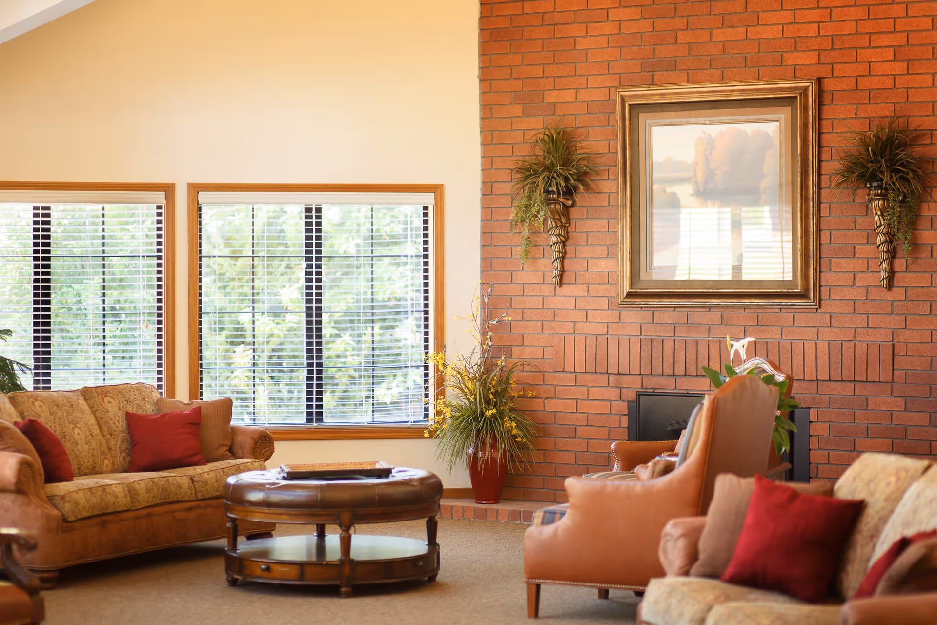 A cozy living room with a brick fireplace wall adorned with a framed landscape painting and two decorative wall sconces with greenery. The room features a patterned sofa with red and brown cushions, two brown armchairs, a round wooden coffee table, and large windows with blinds letting in natural light.