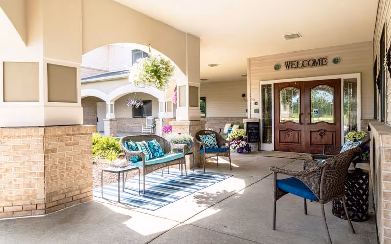 Covered outdoor seating area at the entrance of a building with wicker chairs and a bench with blue cushions, a striped rug, hanging flower baskets, and a wooden double door with a 'WELCOME' sign above it.