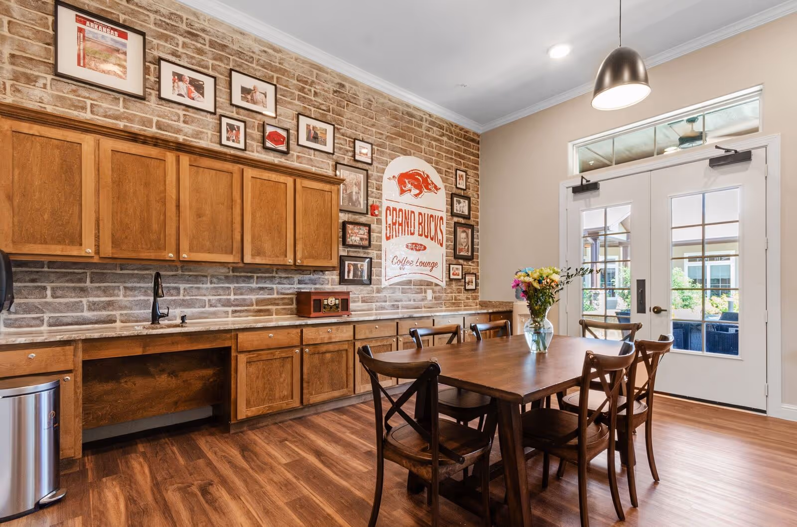 A cozy lounge area with a wooden table and six chairs, a vase with colorful flowers on the table, wooden cabinets along a brick wall, framed pictures and a sign that reads 'Grand Bucks Coffee Lounge', and double glass doors leading outside.