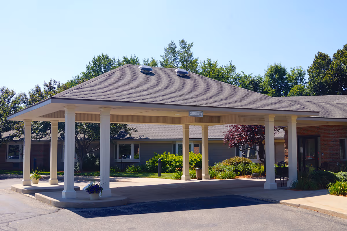 Covered entrance area of Railside Assisted Living Center with white pillars supporting a roof, surrounded by greenery and trees under a clear blue sky.