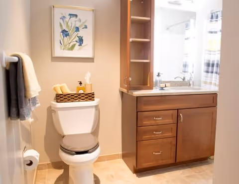 A clean and well-lit bathroom featuring a toilet with a gray seat cover, a basket with towels and toiletries on top, a wooden vanity with a sink and mirror, and a towel rack with hanging towels. A framed floral artwork is hung on the wall above the toilet.