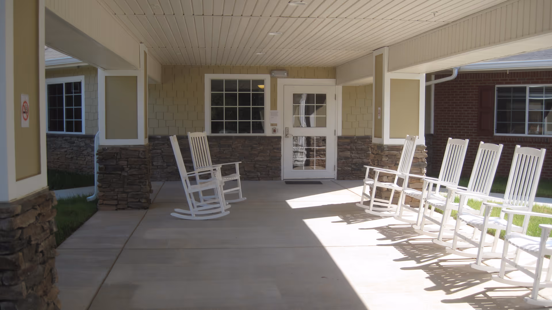 Covered outdoor patio area with white rocking chairs arranged along the sides, stone pillars, and a door with a window leading inside the building.
