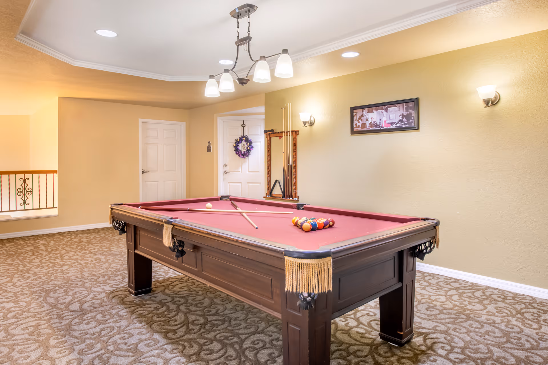 Interior game room with a red-felt pool table, cue sticks, racked balls, chandelier lighting and beige walls.