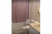 A small bathroom featuring a bathtub with a purple shower curtain, a toilet with a white safety frame around it, a beige countertop with a sink, and a wall-mounted soap dispenser.