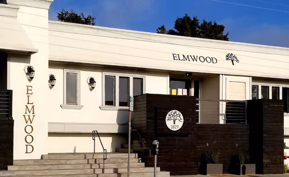 Front exterior view of Elmwood Nursing Rehabilitation Hospital building with steps leading to the entrance, white walls, and black signage with a tree logo and the number 2829.