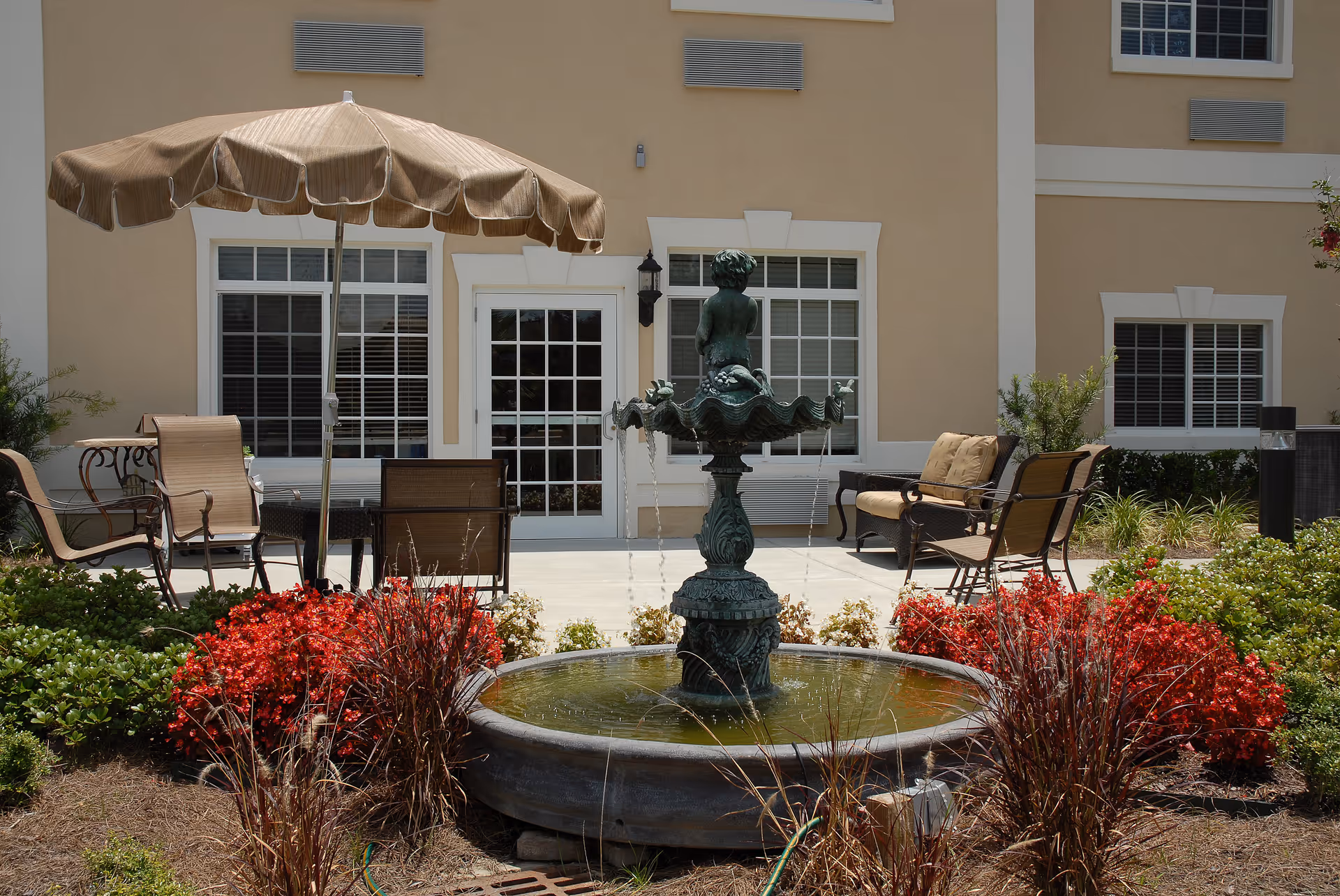 Outdoor patio area with a decorative water fountain featuring a statue of a child sitting on a shell. Surrounding the fountain are red and green plants. The patio has several chairs, a table with a large beige umbrella, and a bench with cushions. The background shows a beige building with white-framed windows and a glass door.