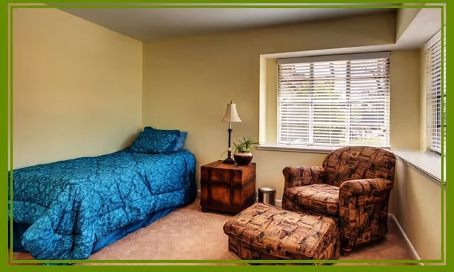 A cozy bedroom featuring a single bed with blue patterned bedding, a wooden nightstand with a lamp and a small plant, and a comfortable armchair with a matching ottoman near a window with white blinds.