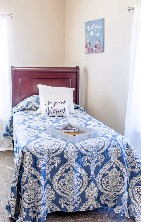 A single wooden bed with a blue patterned bedspread, decorative pillows (one reading "Beyond Blessed"), a tray on the bed, and light-filled pale walls with floral wall art.