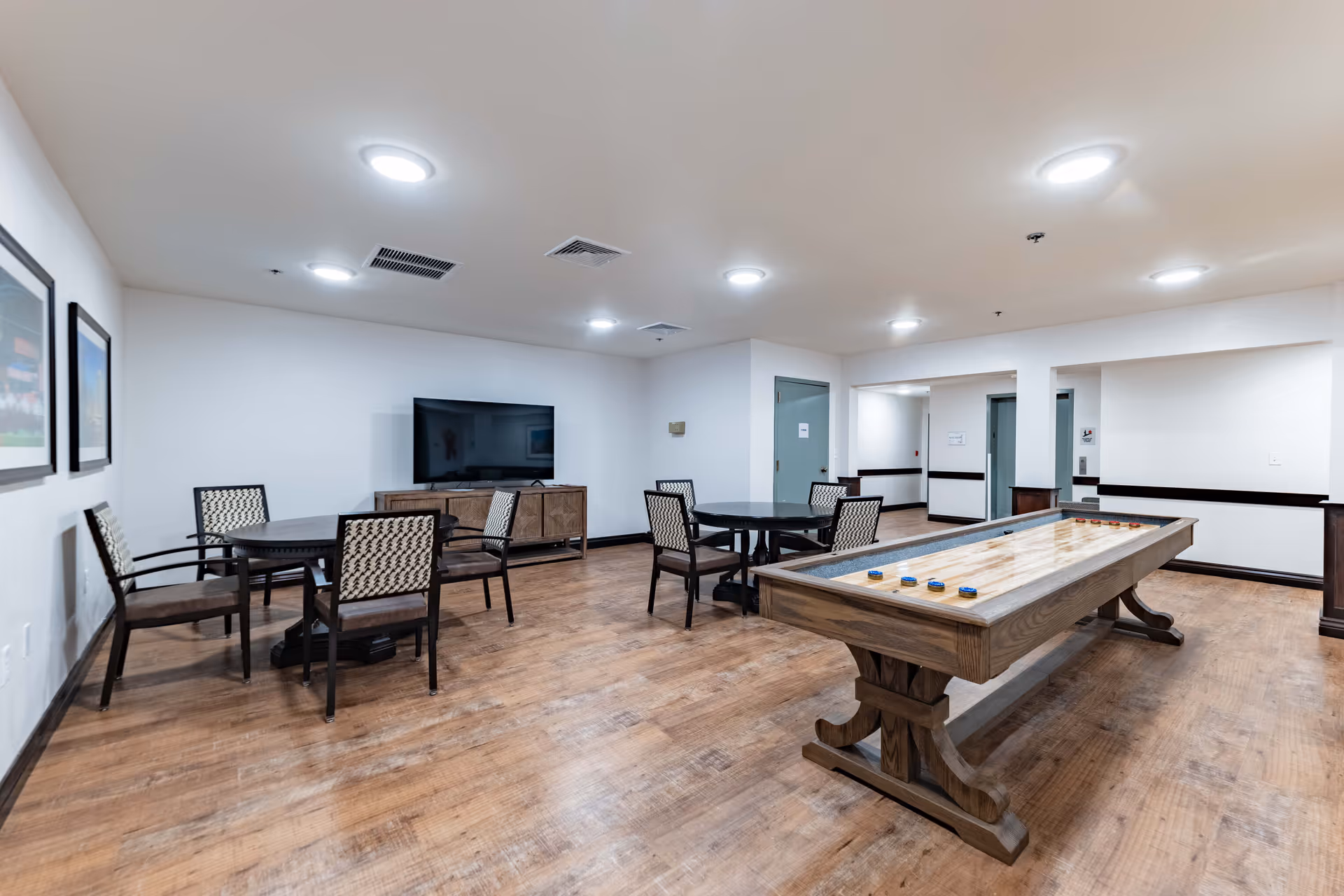 A well-lit senior living common room with a shuffleboard table, round game tables and chairs, and a television.