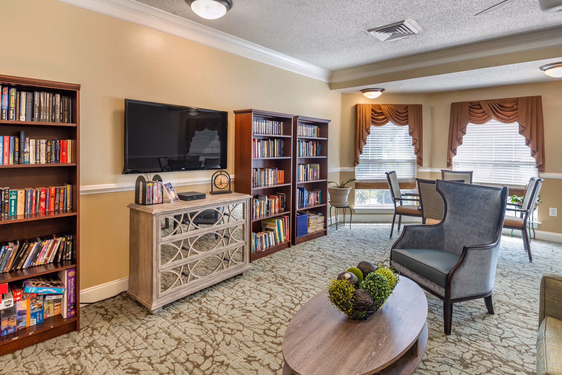 A cozy living room area in a senior living facility featuring a wall-mounted flat-screen TV above a decorative cabinet, two wooden bookshelves filled with books, a comfortable armchair, a wooden coffee table with decorative greenery, and a dining table with chairs near windows with brown valances.