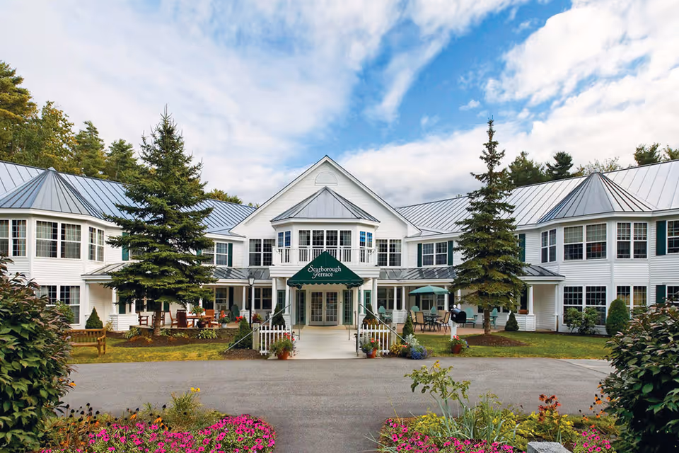 Front exterior view of Scarborough Terrace, a two-story white building with a metal roof, surrounded by trees and landscaped flower beds. There is a green awning over the entrance with the facility name, outdoor seating areas, and a clear sky with some clouds.